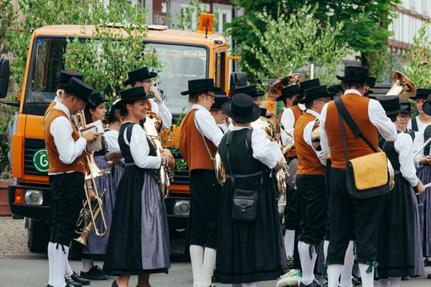 Gruppe von Menschen in traditioneller bayrischer Tracht, die eine Straße entlangmarschieren, Instrumente spielen und Taschen tragen, mit Bäumen, Gebäuden und einem geparkten Fahrzeug im Hintergrund.