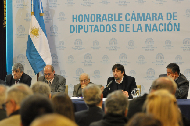 Eine Gruppe von Menschen sitzt an einem Tisch mit einem Krug, Papieren und einem Stift, während eine Person in der Mitte in ein Mikrofon spricht, mit einer weißen und blauen Flagge und einem Hintergrund, auf dem "ehrenwerte Kammer de" steht.
