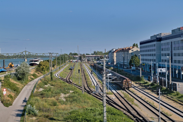 Ein Zug fährt neben städtischer Infrastruktur, einschließlich Gleisen, Strommasten, Kabeln, Gebäuden, Bäumen, Fahrzeugen, einer Brücke, Wasser und einem bewölkten Himmel.