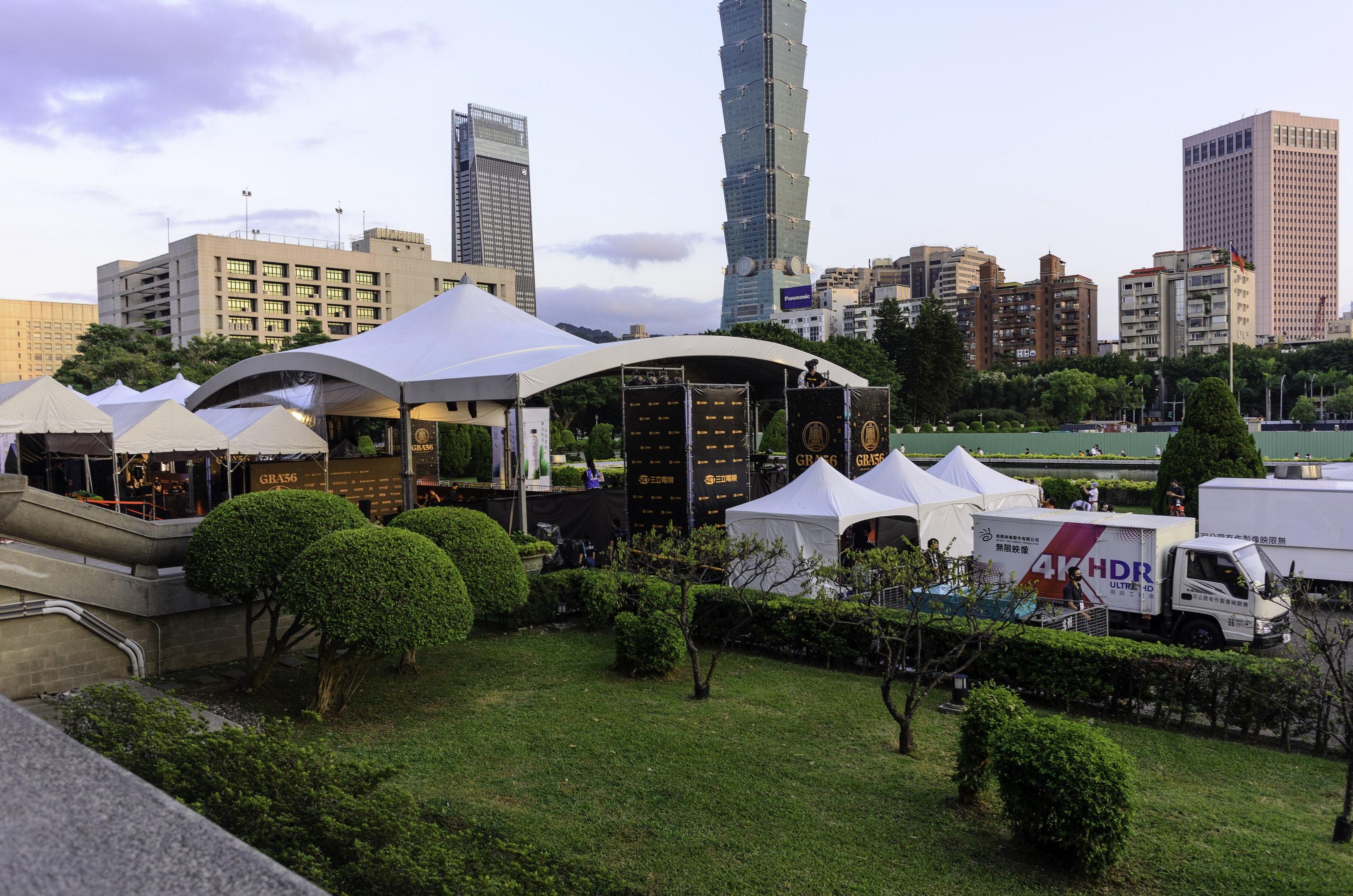 Eine Gruppe von Zelten in einem Park mit Grünflächen, Menschen, Fahrzeugen, Gebäuden und einem klaren Himmel, wahrscheinlich während des Hong Kong International Film Festivals.