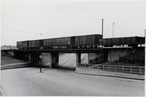 Ein Schwarz-Weiß-Foto eines Zuges, der eine Brücke mit einer Straße darunter, Geländern auf beiden Seiten, Pfählen und Drähten, die entlang der Brücke verlaufen, und einem Turm im Hintergrund überquert.