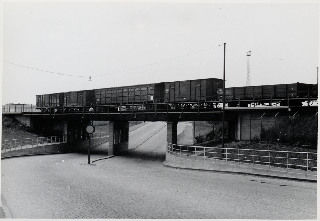 Ein Schwarz-Weiß-Foto eines Zuges, der eine Brücke mit einer Straße darunter, Geländern auf beiden Seiten, Pfählen und Drähten, die entlang der Brücke verlaufen, und einem Turm im Hintergrund überquert.