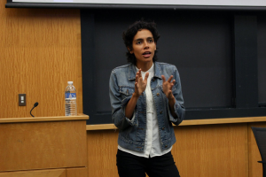 Eine Frau steht an einem Rednerpult mit einem Mikrofon und einer Flasche und hält eine Präsentation vor einem Projektionsschirm, rechts daneben ein Stuhl und eine hölzerne Wand im Hintergrund.