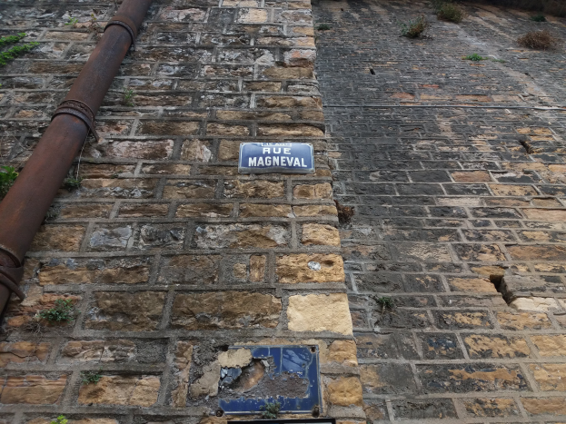 Ein Ziegelgebäude mit einem blauen Schild an der Seite, einer Röhre, die an der Wand entlangläuft, und ein paar Pflanzen, die darum herum wachsen.