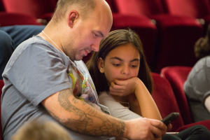 Ein Mann und ein junges Mädchen sitzen in einem Kino, beide schauen auf ein Handy, das der Mann hält.