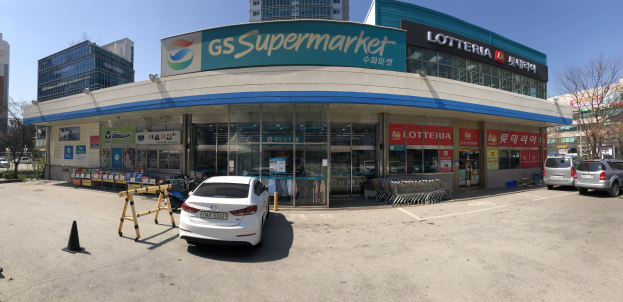 Ein Auto vor einem "GS Supermarkt" parkend mit anderen Fahrzeugen, Verkehrskegeln und verstreuten Gegenständen auf der Straße, Gebäude und Bäume im Hintergrund unter einem klaren blauen Himmel.