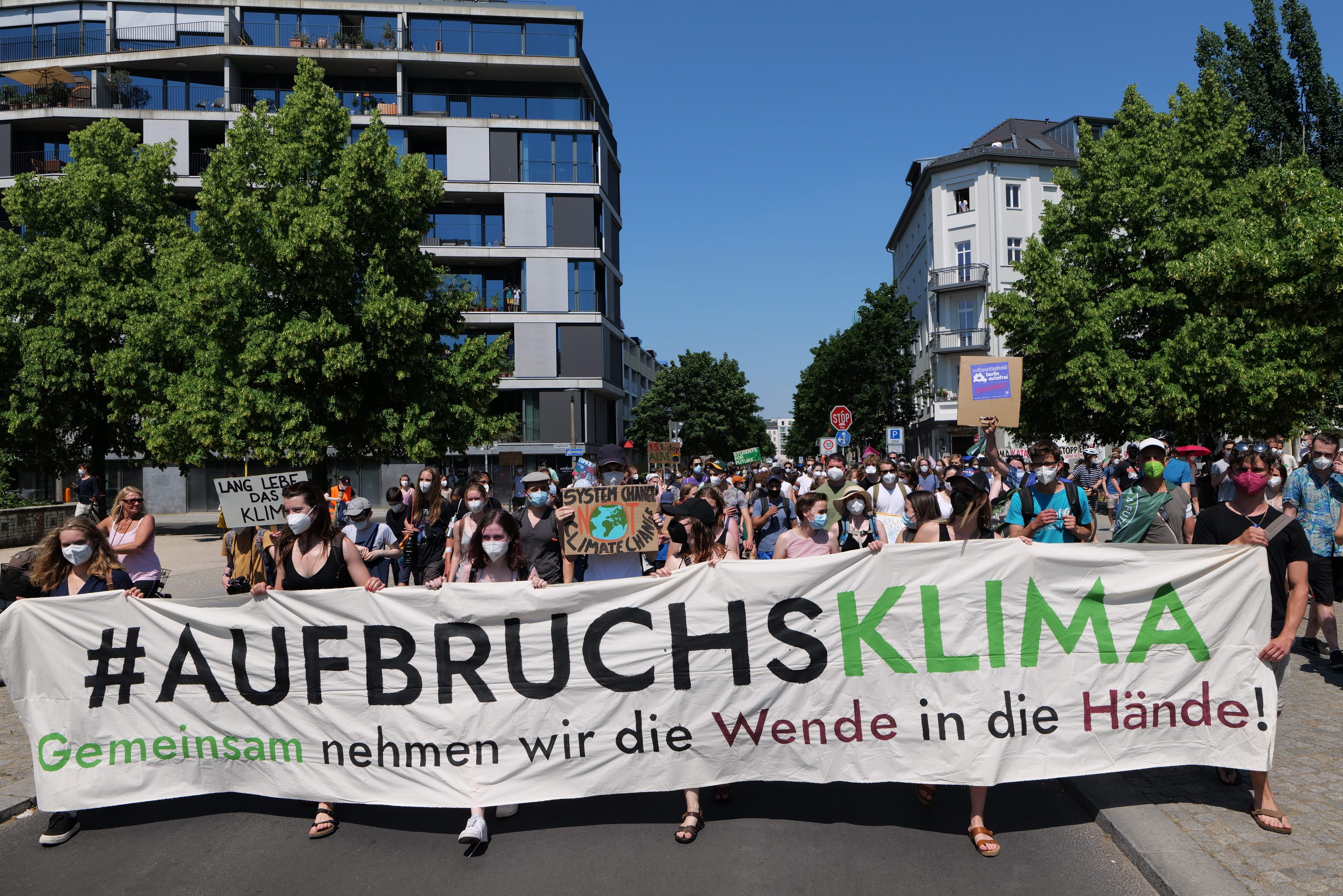 Maskentragende halten ein Banner mit der Aufschrift 'Aufbruchsklima' vor einem Gebäude mit Bäumen und einem klaren Himmel.
