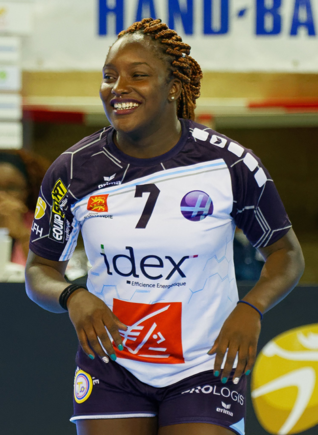 Eine professionelle Handballspielerin in einem lila und weißen Trikot steht lachend auf einem Feld mit einem FIVB-Handball-Weltmeisterschaft-2019-Schild im Hintergrund.
