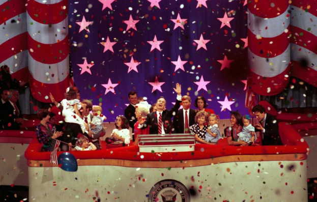 Eine Gruppe von Menschen steht auf einer Bühne mit einem Rednerpult, Ballons und Konfetti, mit Sternen und einem Vorhang im Hintergrund, was auf eine Präsidentenamtseinführung hindeutet.
