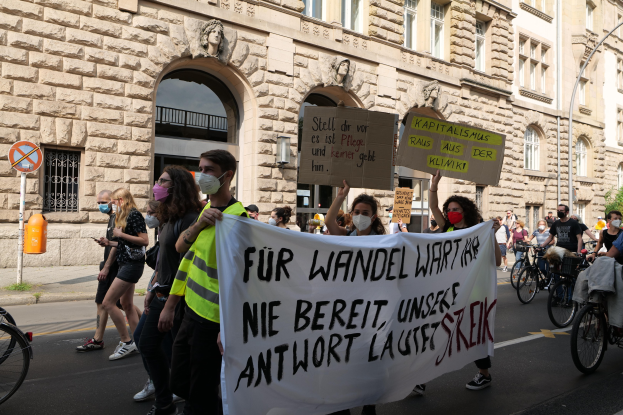 Eine Gruppe von Menschen, die auf einer Demonstration durch die Straße marschieren, einige halten Schilder und andere fahren Fahrräder, mit einem Gebäude im Hintergrund, das Fenster, Bögen, Säulen und Skulpturen aufweist.