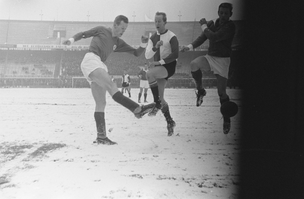 Eine Gruppe von Männern, die Fußball auf einem schneebedeckten Feld spielen, mit einem Stadion im Hintergrund, das von Zuschauern gefüllt ist, unter einem sichtbaren Himmel, dargestellt in Schwarz-Weiß.