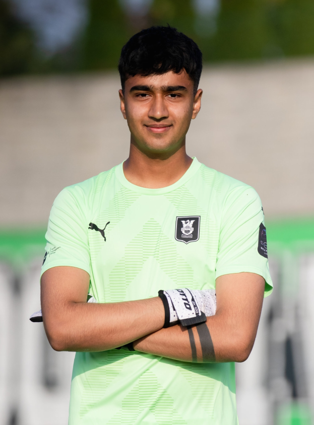 Ein junger Mann in einem grünen T-Shirt steht auf einem Fußballfeld und hat die Arme verschränkt, während er lächelt. Der Hintergrund ist unscharf und suggeriert, dass er im Fokus der Aufnahme ist.