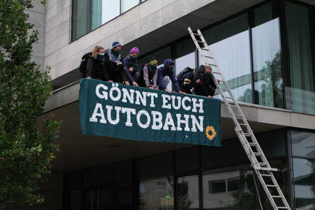 Gruppe von Menschen auf einem Gebäudedach mit einem Banner, einem Baum auf der linken Seite und Glasfenstern im Hintergrund, die scheinbar für die deutsche Regierung protestieren.