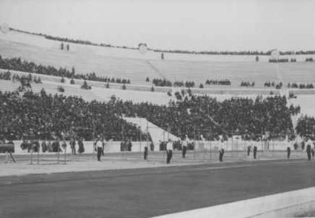 Schwarzes und weißes Foto eines vollen Stadions während eines Ereignisses, mit einer Straße unten, Bäumen und Himmel im Hintergrund, wahrscheinlich die Eröffnungszeremonie der Olympischen Spiele 1936 im Londoner Olympiastadion.