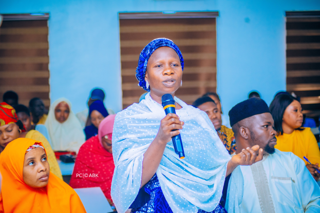 Eine Frau in einem blauen Kleid spricht vor einer sitzenden Gruppe, einige halten Stifte, in einer Kirche, wahrscheinlich über Frauenrechte in Nigeria.