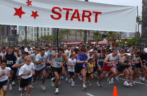Gruppe von Menschen beim Marathon mit einem Verkehrskegel im Vordergrund und einer Fahne im Hintergrund, umgeben von Bäumen, Laternenmasten, Gebäuden und einem klaren blauen Himmel.
