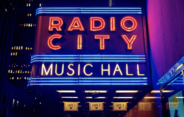Das erleuchtete Radio City Music Hall in New York City, umgeben von hohen Gebäuden, mit einem "Radio City"-Schild und zusätzlichen Schildern auf der rechten Seite.