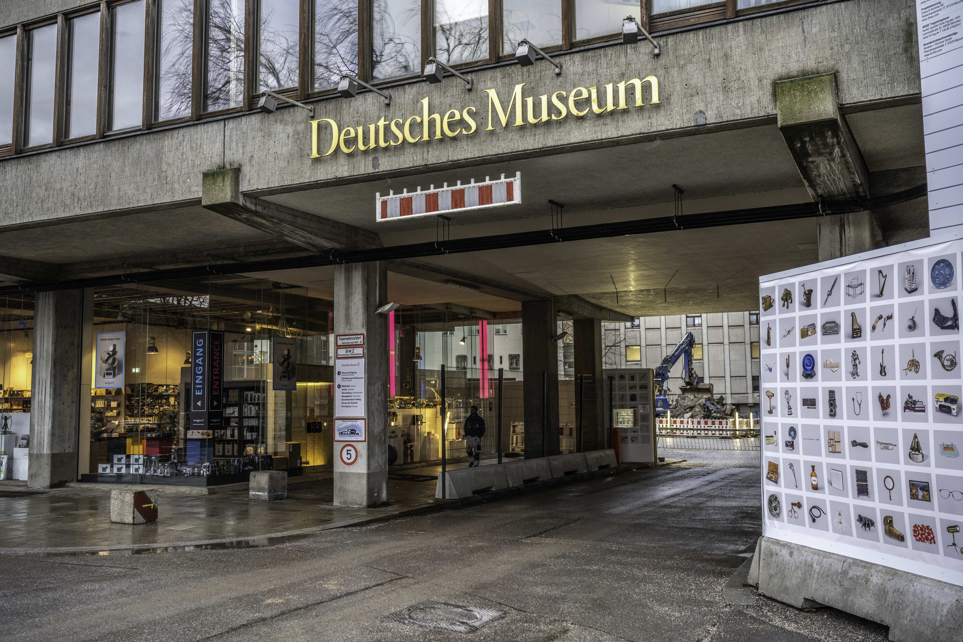Großes gläsernes Gebäude des Deutschen Museums in Berlin mit einem Namensschild und einer informativen Anzeigetafel auf der rechten Seite, vor einem Hintergrund aus anderen Gebäuden, Bäumen und Himmel.