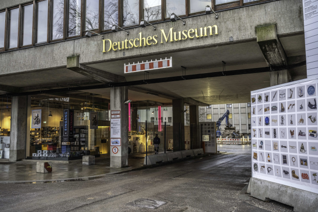 Großes gläsernes Gebäude des Deutschen Museums in Berlin mit einem Namensschild und einer informativen Anzeigetafel auf der rechten Seite, vor einem Hintergrund aus anderen Gebäuden, Bäumen und Himmel.