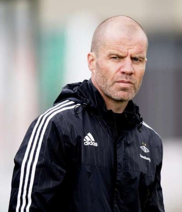 Baldiger Trainer in einer blauen Jacke mit weißen Streifen und weißem Kragen, der auf einem Fußballfeld steht und die Hände verschränkt hält und einen ernsten Gesichtsausdruck hat.