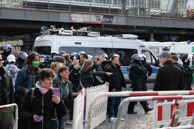 Eine Gruppe von Menschen vor Polizeifahrzeugen mit Absperrungen im Vordergrund während einer Demonstration in Berlin, Deutschland.