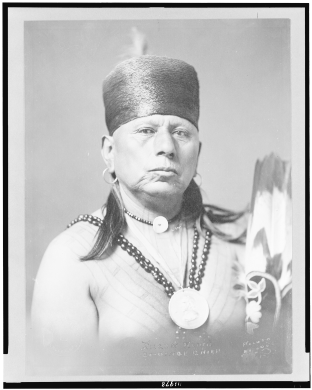 Ein altes Schwarz-Weiß-Foto eines indianischen Mannes mit langen dunklen Haaren, der eine Mütze und traditionelle indianische Kleidung trägt und direkt in die Kamera schaut.