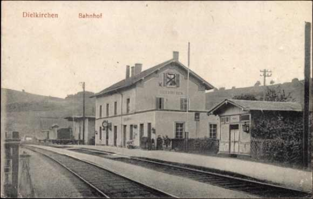 Schwarzes und weißes Foto einer Bahnstation in Dielkirchen, Deutschland, mit einem Zug auf den Gleisen, umgeben von Häusern, Strommasten, Bäumen, Menschen und Bergen sowie einem klaren Himmel im Hintergrund.