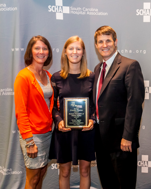 Drei Personen, zwei Frauen und ein Mann, stehen zusammen. Der Mann trägt einen schwarzen Anzug, eine Frau trägt ein orange-schwarzes Kleid und die andere Frau trägt ein schwarzes Kleid und hält einen Award in der Hand. Sie stehen vor einem grauen Hintergrund.