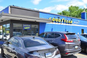 Ein Goodyear Select Autohaus mit mehreren parkenden Autos, das ein Gebäude mit Text und einen Hintergrund aus Bäumen unter einem bewölkten Himmel zeigt.