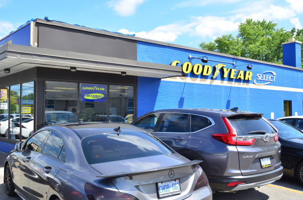 Ein Goodyear Select Autohaus mit mehreren parkenden Autos, das ein Gebäude mit Text und einen Hintergrund aus Bäumen unter einem bewölkten Himmel zeigt.