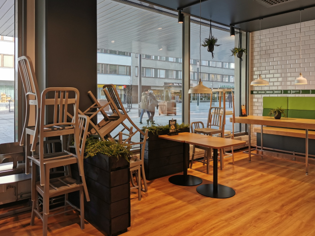 Ein Restaurant-Innenraum mit Holztischen und -stühlen in der Nähe eines großen Fensters, das auf eine Berliner Straße mit Passanten, Gebäuden und städtischen Elementen blickt.