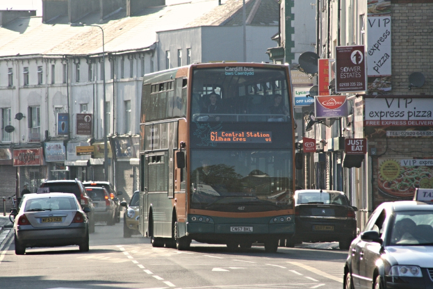 Eine Straße mit Autos und einem Bus im Vordergrund, mit Gebäuden mit Wänden, Fenstern, Tellern und Dächern im Hintergrund, geschmückt mit Plakaten und Bannern, und einem Laternenmast.