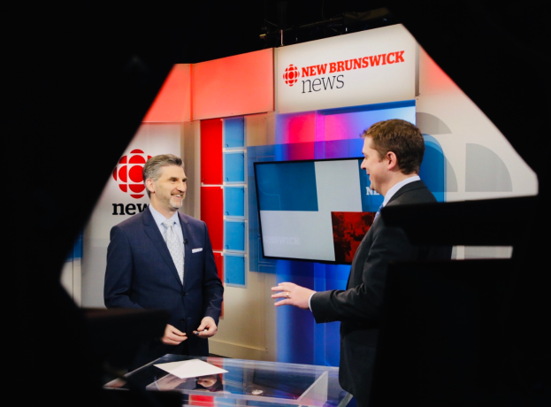 Zwei Männer in Blazern und Krawatten vor einem Glastisch mit Papieren, im Hintergrund ein Fernseher mit der Aufschrift "New Brunswick News".