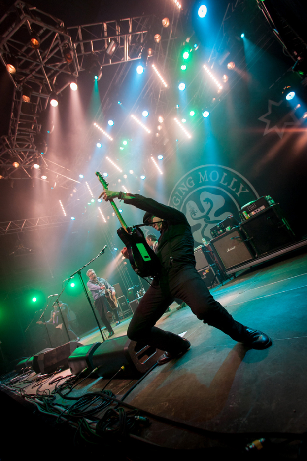 Ein musikalisches Konzert mit drei Künstlern auf der Bühne, zwei spielen Gitarre mit Mikrofonen davor, ein Hintergrund mit dem Wort "MOLLEY" und zwei Schlangensymbole sowie Scheinwerfer darüber.