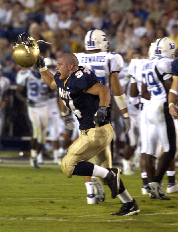 Ein Fußballspieler in dunkler Uniform rennt mit dem Ball, umgeben von Teamkollegen in Sportkleidung und Helmen auf dem Feld, während Zuschauer auf Stühlen im Hintergrund sitzen.