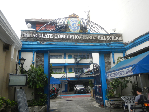 Eingang zur Unbefleckten Empfängnis Pfarrschule mit einem Bogen mit Text, umgeben von Gebäuden, Grünflächen, Fahrzeugen und Menschen unter einem sichtbaren Himmel.