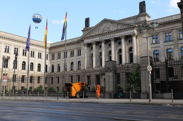 Großes modernes Regierungsgebäude mit zahlreichen Fenstern, umgeben von Straßeninfrastruktur, Fußgängern und Bäumen, unter einem bewölkten Himmel.