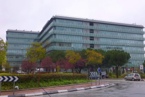 Großes modernes Gebäude mit zahlreichen Fenstern, als Europäisches Zentralbank-Hauptquartier bezeichnet, umgeben von Grünflächen, Straßeninfrastruktur und Fahrzeugen unter einem bewölkten Himmel.