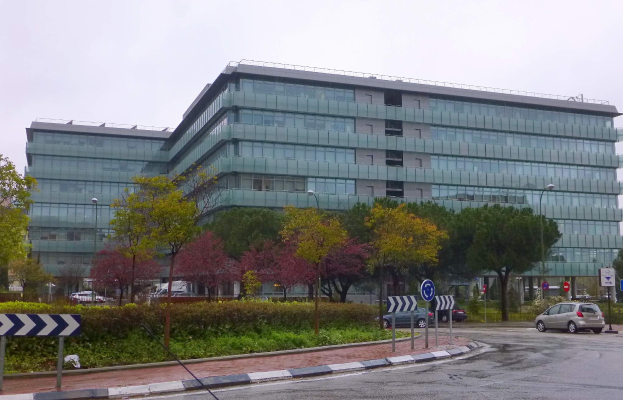 Großes modernes Gebäude mit zahlreichen Fenstern, als Europäisches Zentralbank-Hauptquartier bezeichnet, umgeben von Grünflächen, Straßeninfrastruktur und Fahrzeugen unter einem bewölkten Himmel.