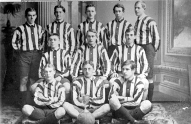 Ein Schwarz-Weiß-Foto einer Fußballmannschaft, mit einigen Spielern, die stehen und andere auf dem Boden sitzen, einem Pokal in der Mitte, Säulen und einer Wand im Hintergrund und Text oben und unten auf dem Foto.