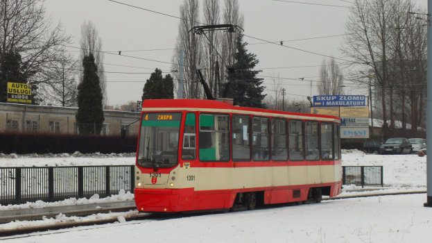 Ein roter und weißer Zug fährt auf schneebedeckten Schienen, umgeben von einem Metallzaun, einem Pfahl, Bäumen, Autos, Gebäuden mit Fenstern, Schildern, Strommasten mit Drähten und einem bewölkten Himmel.
