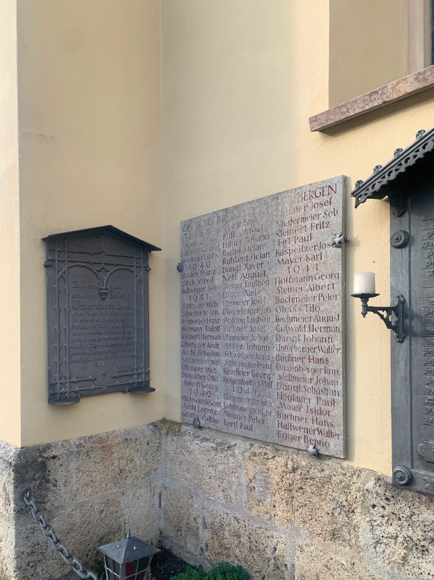 Ein Gebäude mit einer Holocaust-Gedenktafel, einem Fenster, einer Tür, einer Außenleuchte, einer Kette und einigen Pflanzen.