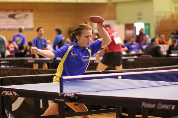 Eine Frau mit einem Schläger in der Hand wird in den Vordergrund gestellt und spielt Tischtennis, während im Hintergrund einige Personen sitzen und stehen und ein Banner an einer Wand befestigt ist.