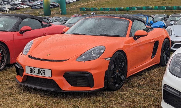 Ein Porsche 718 Boxster GTS auf dem Goodwood Festival of Speed zusammen mit anderen Autos, einem Banner, Bäumen und einem klaren blauen Himmel im Hintergrund.