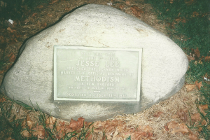 Ein großer Felsen mit einer Tafel, auf der 'Jesse Lee Methodism' steht, in einer grasbewachsenen Fläche umgeben von trockenen Blättern und Gras.