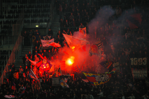 Eine große Menschenmenge in einem Stadion mit Fahnen und Bannern, beleuchtet von Scheinwerfern und umgeben von Stufen und Geländern, mit einem Feuer in der Mitte.
