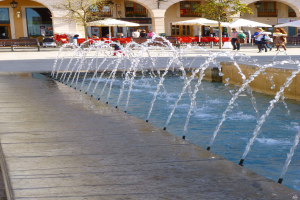 Ein Stadtplatz mit einem zentralen Brunnen, Menschen, die herumlaufen, Bänke, Bäume, Sonnenschirme, Tische, Stühle und ein Gebäude mit Bögen, Säulen, Fenstern und Lichtern im Hintergrund.