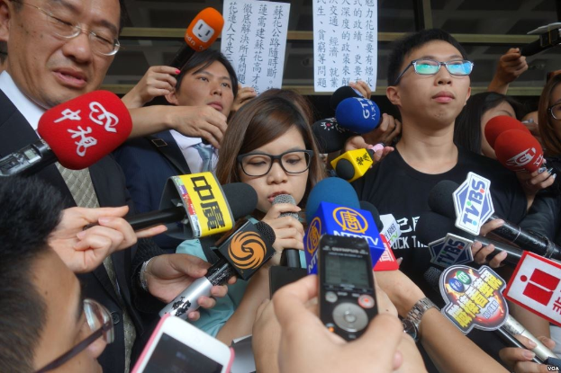 A group of people engaged in a discussion outside, surrounded by reporters holding microphones, phones, and papers, with a glass wall in the background.