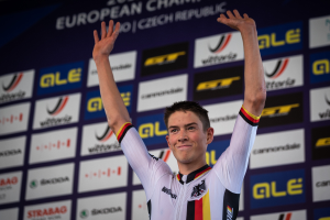 Ein Mann in buntem Radfahrer-Outfit steht auf einem Podium mit erhobenen Händen, mit einem "Giro d'Italia"-Schild im Hintergrund.