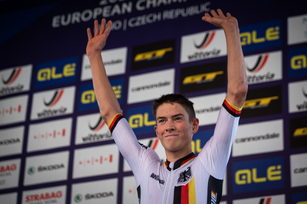 Ein Mann in buntem Radfahrer-Outfit steht auf einem Podium mit erhobenen Händen, mit einem "Giro d'Italia"-Schild im Hintergrund.